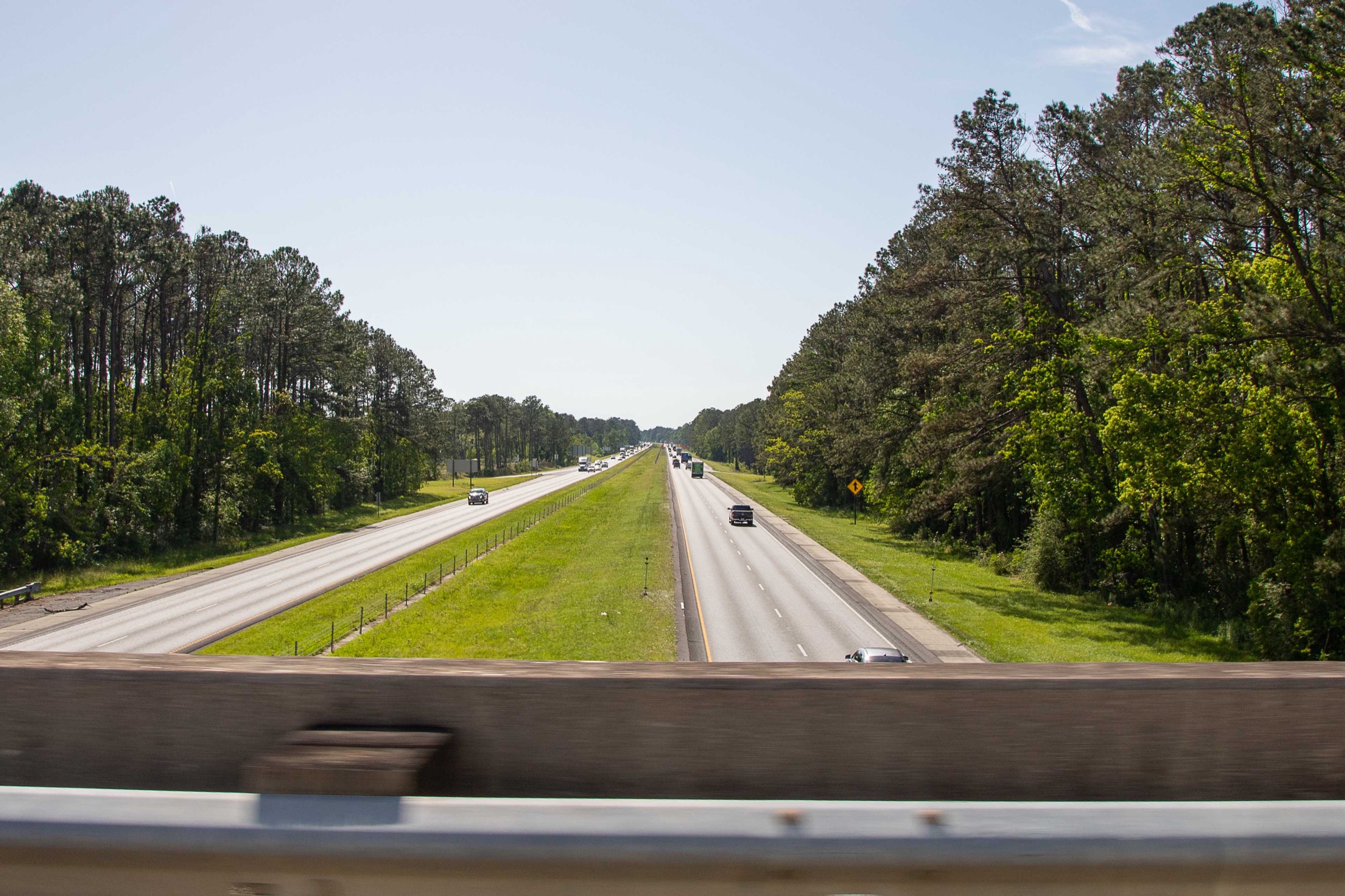 Looking east down Interstate 12 from an overpass — twin dual-carriageway running between pine woods, the corridor the Managed Lanes Conversion adds HOV capacity to
