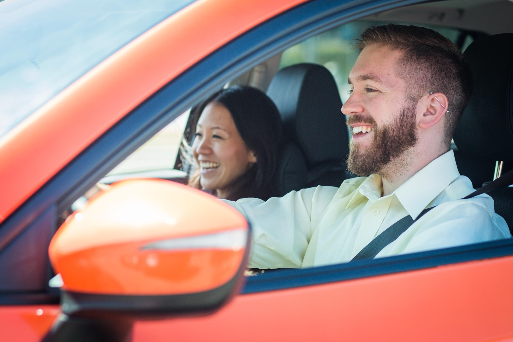 A driver and passenger laughing together in the front seats of an orange car, shot through the open driver's window