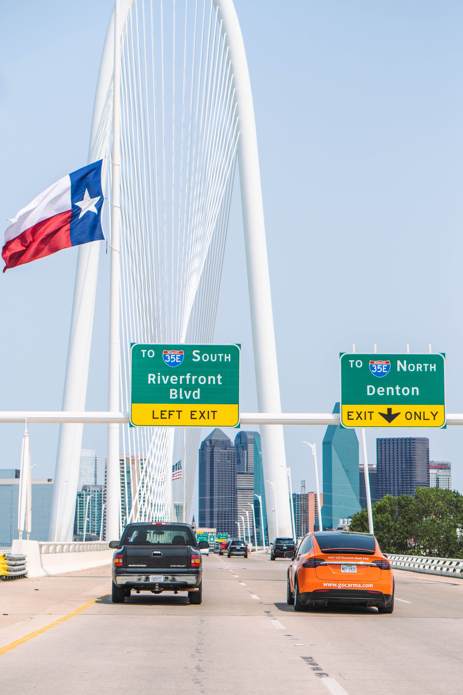 The orange GoCarma Tesla driving north across the Margaret Hunt Hill Bridge into downtown Dallas, Texas state flag flying alongside