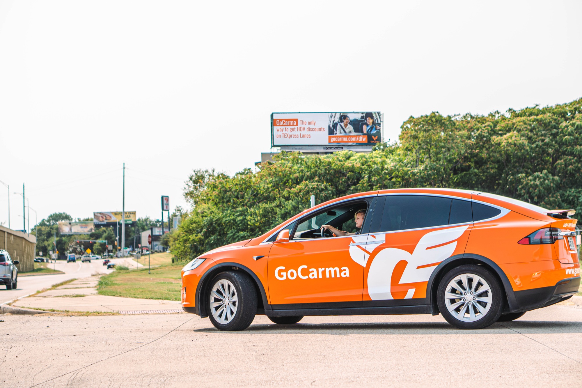 The orange GoCarma Tesla Model X driving past a Dallas billboard reading 'GoCarma — The only way to get HOV discounts on TEXpress Lanes'