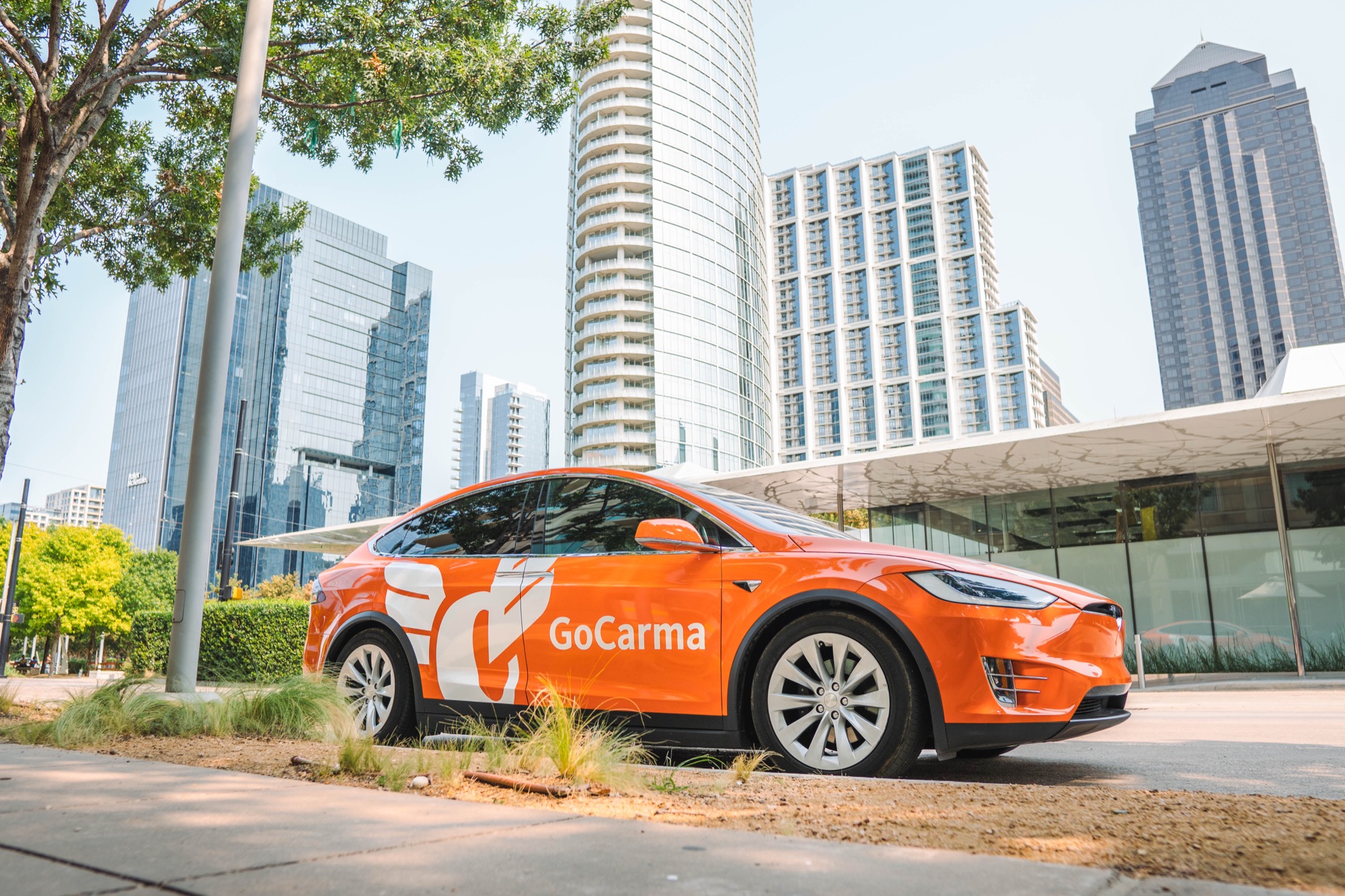 The orange GoCarma Tesla Model X parked beside high-rise buildings in central Dallas with the downtown skyline behind