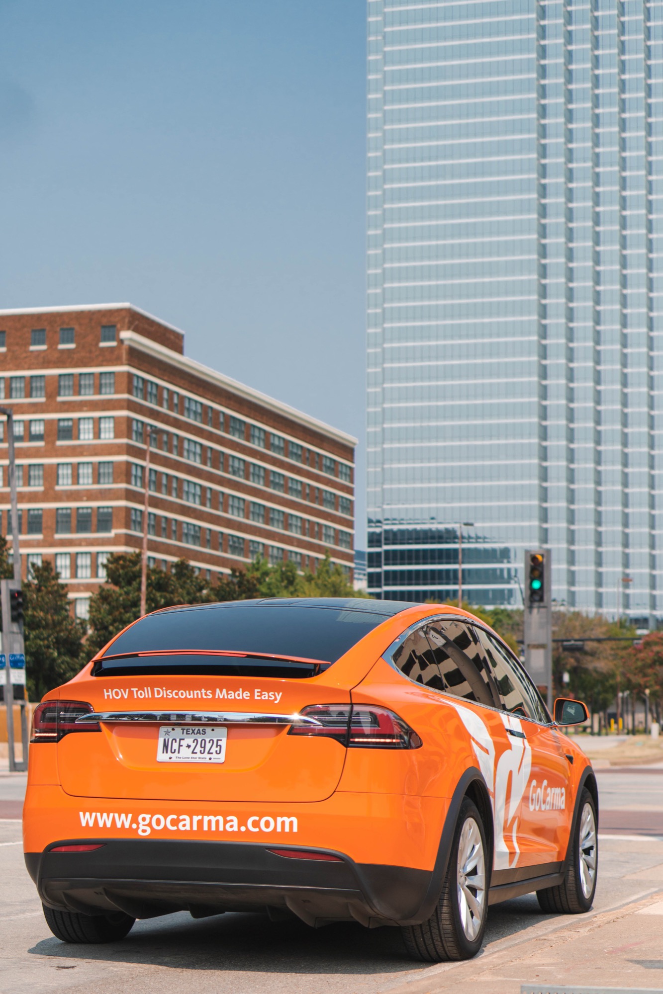 The orange GoCarma Tesla Model X parked at a downtown Dallas intersection, 'HOV Toll Discounts Made Easy' decal visible on the rear with the Dallas skyline behind